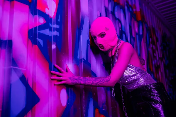 Anonima donna tatuata in passamontagna toccando parete con graffiti colorati e guardando la fotocamera in luce viola al neon — Foto stock