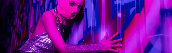Сексуальная женщина в розовой балаклаве смотрит в камеру, прикасаясь к красочным граффити в фиолетовом свете, баннер — стоковое фото