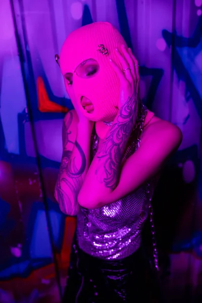 Passionate tattooed woman touching pink balaclava while standing with closed eyes near colorful graffiti in purple light — Stock Photo