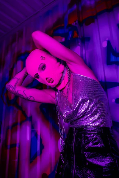 Низкий угол обзора провокационной женщины в балаклаве и металлическом топе, смотрящей на камеру в фиолетовом свете рядом с красочными граффити — стоковое фото