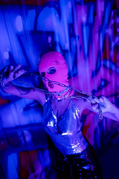 High angle view of provocative woman in balaclava looking at camera while posing with silver chain near graffiti in blue neon light — Stock Photo