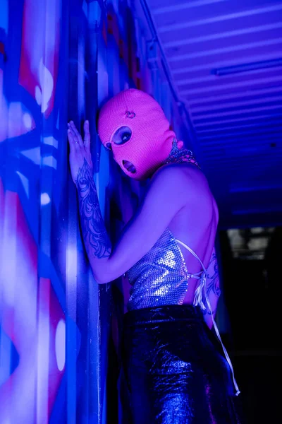 Femme sexy en cagoule et haut métallique regardant la caméra près du mur avec des graffitis en éclairage bleu et violet — Photo de stock