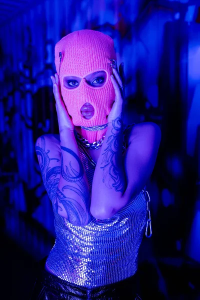 Femme tatouée en cagoule et dessus métallique avec des colliers en argent posant avec les mains près du visage en lumière bleue et violette — Stock Photo