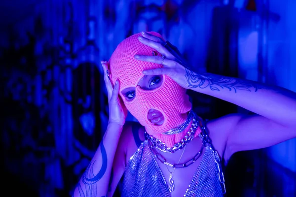 Femme tatouée passionnée dans des chaînes de cou en argent regardant la caméra tout en touchant cagoule tricotée dans la lumière bleue et violette — Photo de stock