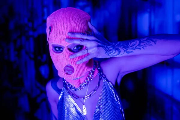 Provokante Frau in Sturmhaube und metallischem Oberteil mit Halsketten, die mit händennahem Gesicht in blauer und lila Beleuchtung posiert — Stockfoto