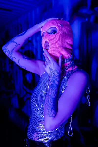 Mulher tatuada em top brilhante e balaclava de mãos dadas perto do rosto enquanto olha para a câmera em luz azul e roxa — Fotografia de Stock