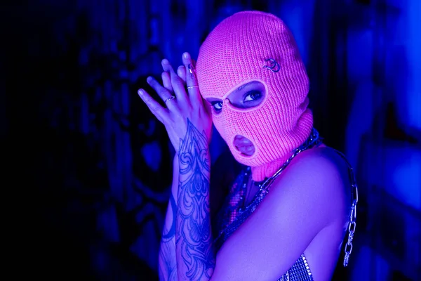 Young tattooed woman in balaclava and silver neck chains looking at camera near wall with graffiti in blue and purple light — Stock Photo