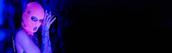 Mulher tatuada anônima em balaclava olhando para a câmera em luz azul e roxa perto do espaço de cópia preta, banner — Fotografia de Stock