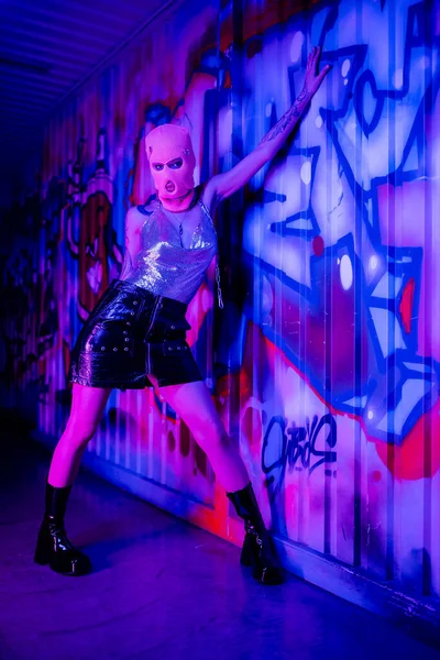 Full length of sexy woman in balaclava and black leather skirt posing near colorful graffiti in blue and purple light — Stock Photo