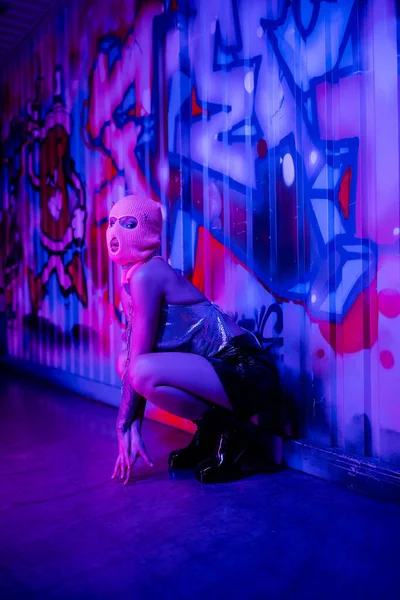 Полная длина женщины в стильной одежде и балаклаве сидя у стены с граффити в синем и фиолетовом освещении — стоковое фото
