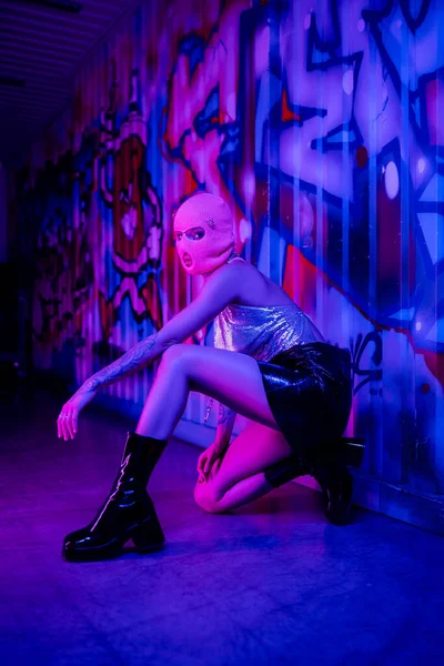 Pleine longueur de femme passionnée en cagoule et jupe en cuir avec des bottes posant près du mur avec des graffitis en lumière bleue et violette — Photo de stock