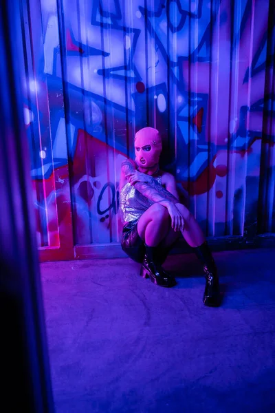 Longitud completa de la mujer sexy en pasamontañas y botas de cuero mirando hacia otro lado mientras está sentado cerca de graffiti colorido en luz azul y púrpura - foto de stock