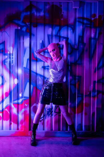 Pleine longueur de la femme en tenue glamour et cagoule posant avec les mains derrière la tête près des graffitis dans la lumière bleue et violette — Photo de stock
