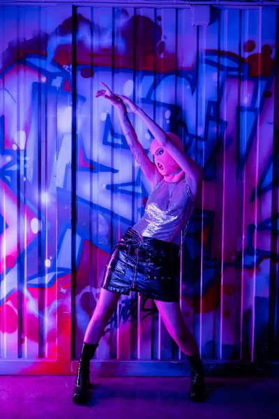 Full length of passionate woman in black leather skirt and balaclava standing with raised hands near colorful graffiti in blue and purple lighting — Stock Photo