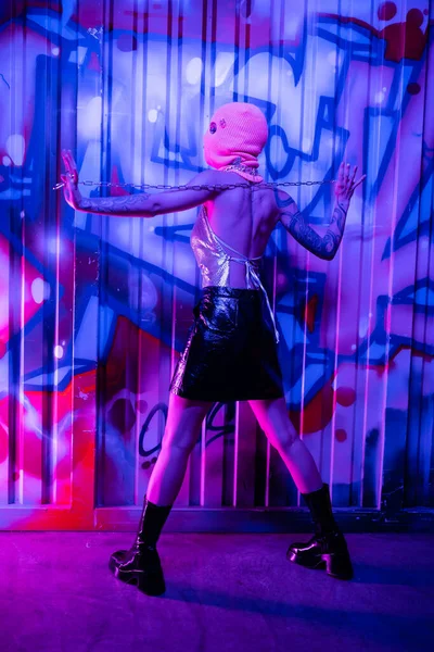 Vue arrière de femme provocatrice en bottes noires et cagoule posant avec chaîne argentée près de graffitis colorés en néon bleu — Photo de stock