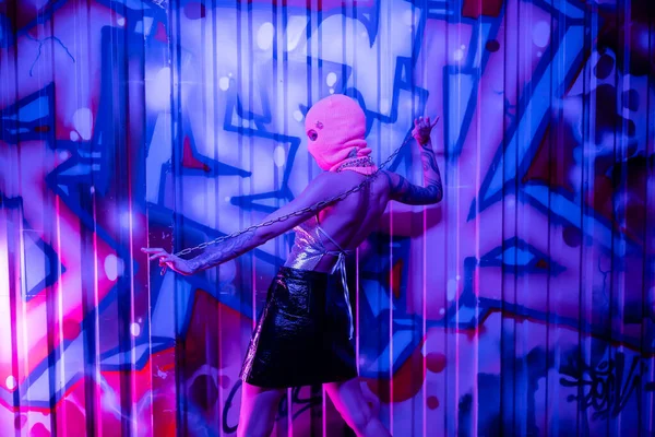 Боковой вид провокационной женщины в черной кожаной юбке и балаклаве, позирующей с цепью рядом с красочными граффити в синем и фиолетовом свете — стоковое фото