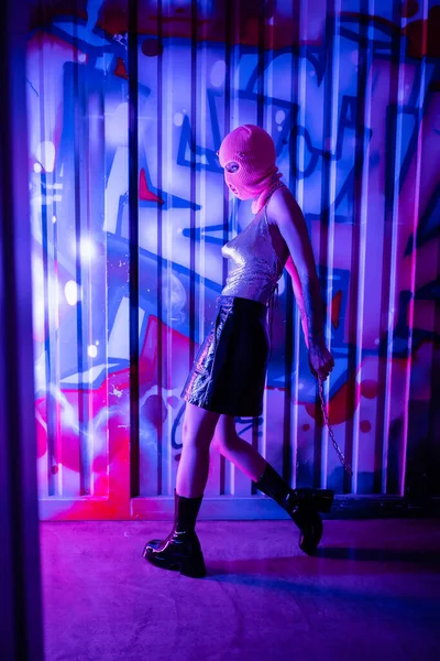 Vista laterale di donna sexy in abito elegante e passamontagna in piedi con catena vicino alla parete con graffiti in luce blu e viola — Foto stock
