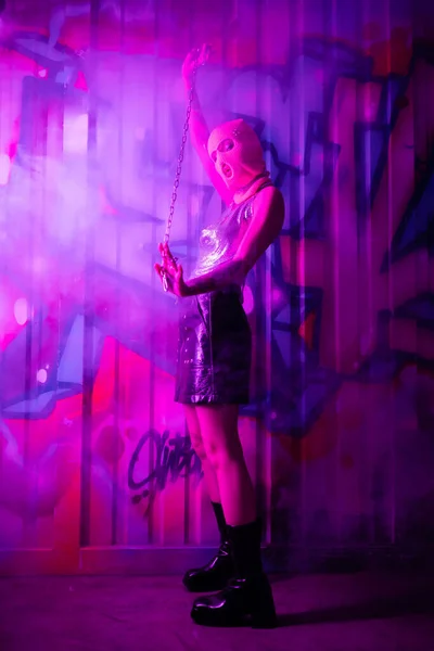 Longitud completa de la mujer en pasamontañas y botas de cuero posando con cadena cerca de graffiti colorido en luz púrpura con humo - foto de stock