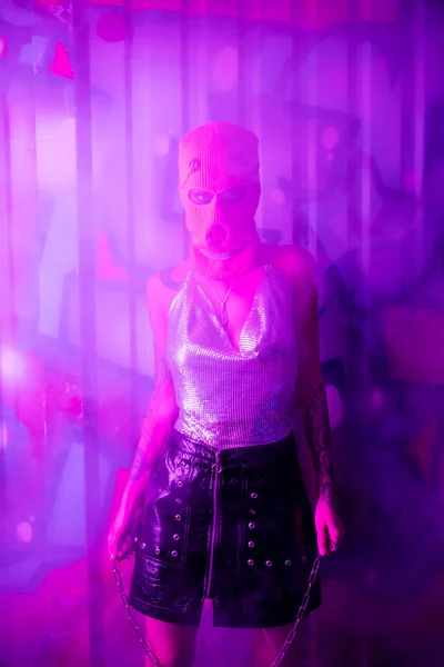 Sexy Frau in Sturmhaube und silbernem Top mit Halskette und Blick auf Kamera in Wandnähe mit Graffiti in lila Licht mit Rauch — Stockfoto