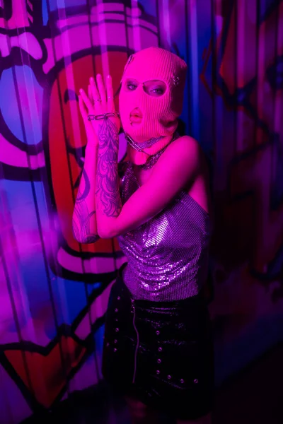Passionate woman in pink balaclava holding chain and looking at camera near colorful graffiti in purple neon light — Stock Photo