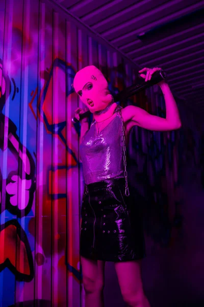 Стильна жінка в Балаклаві і металевому верхньому позуванні з бейсбольною битою біля стіни з графіті в фіолетовому неоновому світлі — стокове фото