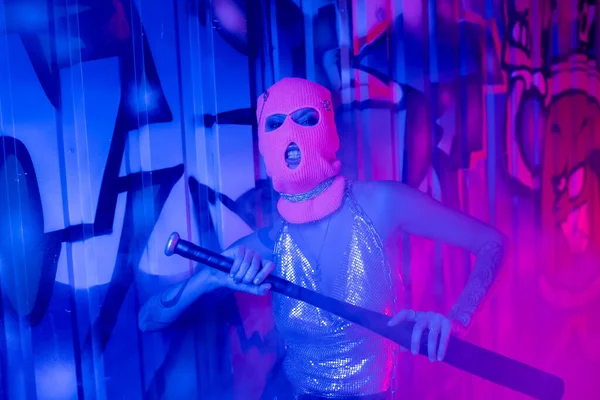 Femme provocatrice en cagoule et dessus argenté grimacant avec batte de baseball près de graffitis dans la lumière bleue et violette — Photo de stock