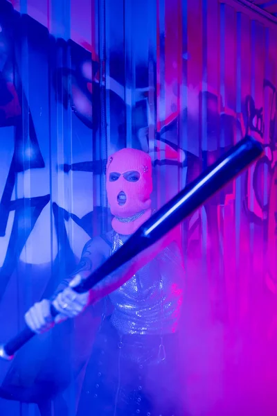 Агрессивная женщина в балаклаве гримасит, держа бейсбольную биту возле граффити в синем и фиолетовом свете с дымом — стоковое фото