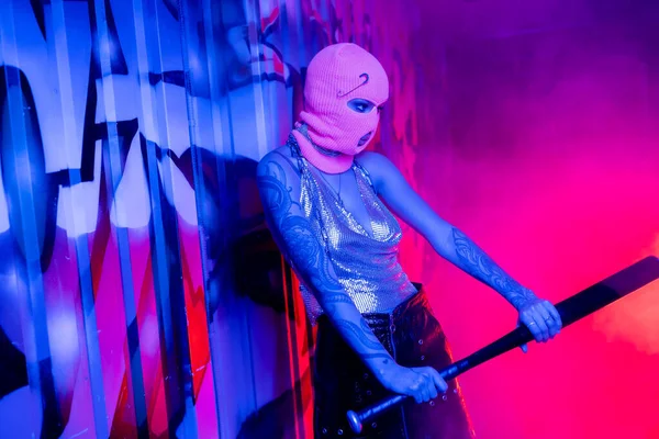 Опасная женщина в балаклаве и металлическом топе с бейсбольной битой возле граффити в синем неоновом свете рядом с розовым дымом — стоковое фото