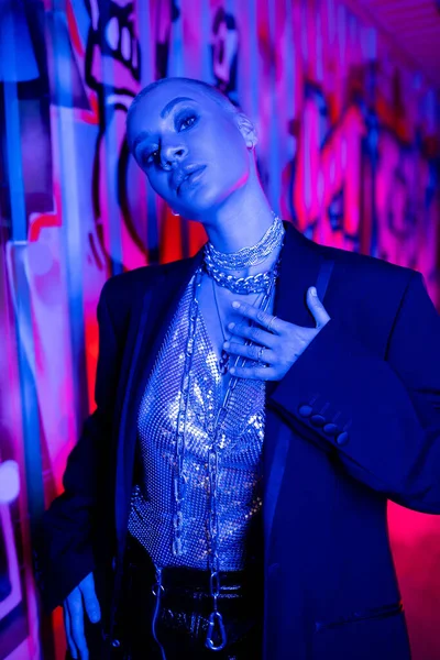 Seductive woman in black blazer and shiny top with silver neck chains touching chest near colorful graffiti in blue and purple lighting — Stock Photo