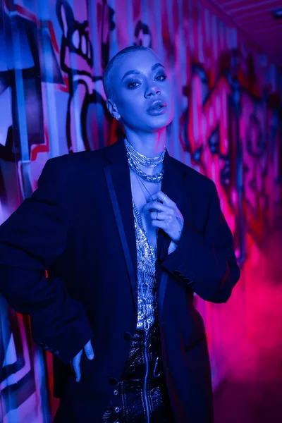 Модная коротковолосая женщина в черном блейзере и серебряных ожерельях, смотрящая на камеру рядом с граффити в синем свете с розовым дымом — стоковое фото