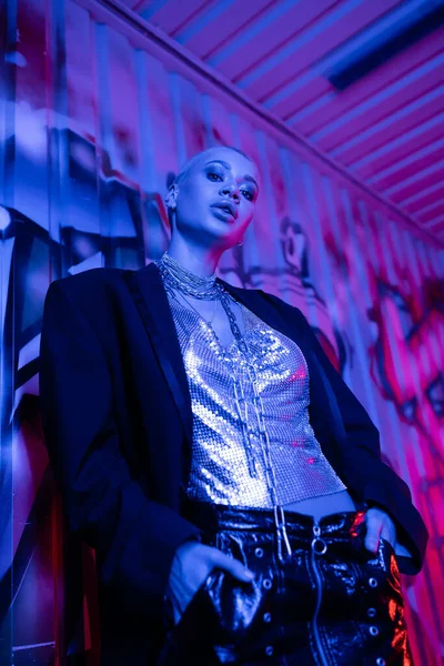 Visão de baixo ângulo da mulher sexy no topo metálico e casaco preto posando com as mãos em bolsos perto de grafite em luz azul e roxa — Fotografia de Stock