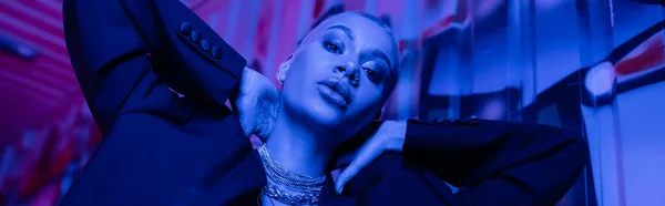 Tiefansicht einer verführerischen Frau im schwarzen Blazer und silbernen Halsketten, die die Hände hinter dem Hals in blauem Neonlicht hält, Banner — Stockfoto