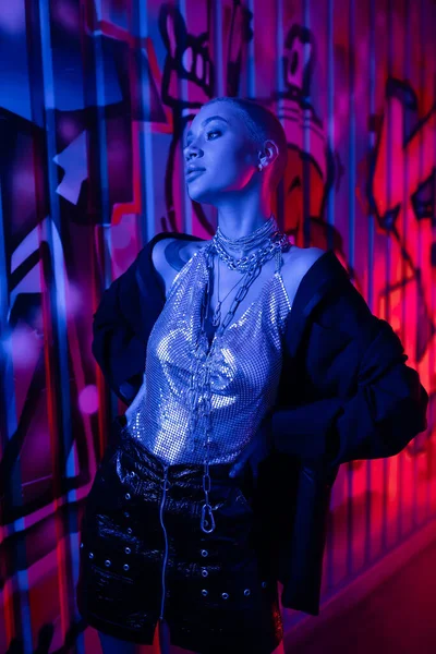 Блискуча жінка в металевому верхньому і срібному ланцюжках шиї позує руками на талії біля барвистого графіті в блакитному неоновому світлі — стокове фото