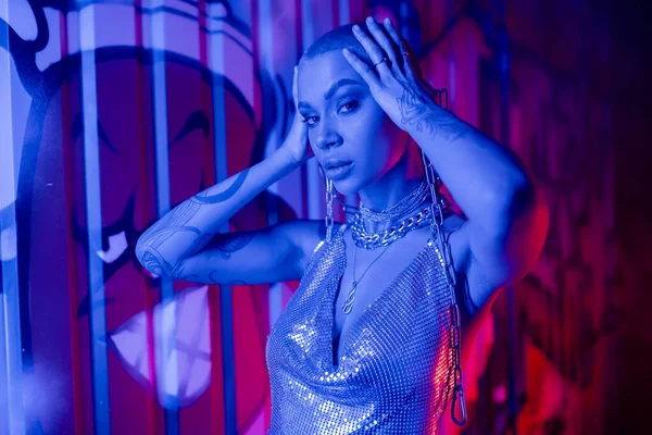 Mujer tatuada extravagante en la parte superior brillante cogida de la mano cerca de la cabeza mientras mira la cámara cerca de la pared con graffiti en la iluminación azul - foto de stock