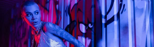 Sexy donna in top metallico e catene collo guardando lontano vicino graffiti colorati in luce blu al neon, banner — Foto stock