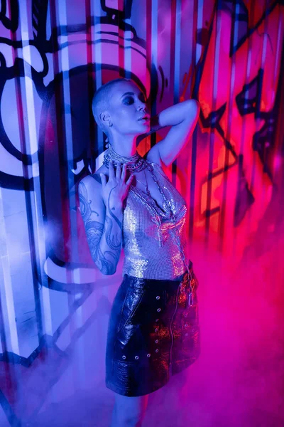 Femme tatouée passionnée en haut métallique et jupe en cuir noir debout près de graffitis colorés dans la lumière bleue et rose avec de la fumée — Photo de stock