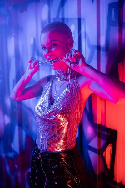 Провокационная женщина в металлическом топе кусает серебряную цепочку и смотрит на камеру в голубом и розовом свете рядом с красочными граффити — стоковое фото