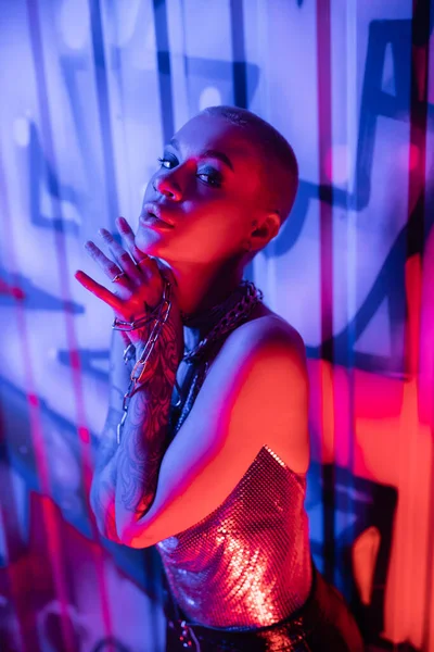 Tattooed woman looking at camera while posing with silver chains near wall with graffiti in blue and pink lighting — Stock Photo
