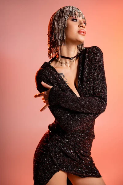 Femme tatouée de luxe en perruque bijoux et robe lurex noir regardant loin sur fond rose corail — Photo de stock