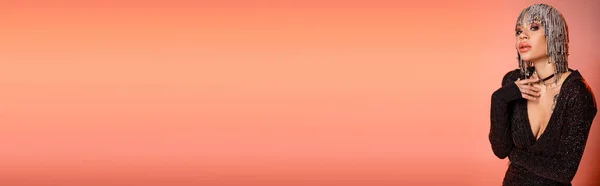 Donna alla moda e sexy in copricapo metallico e abito nero in posa con mano vicino al petto su sfondo rosa corallo, banner — Foto stock