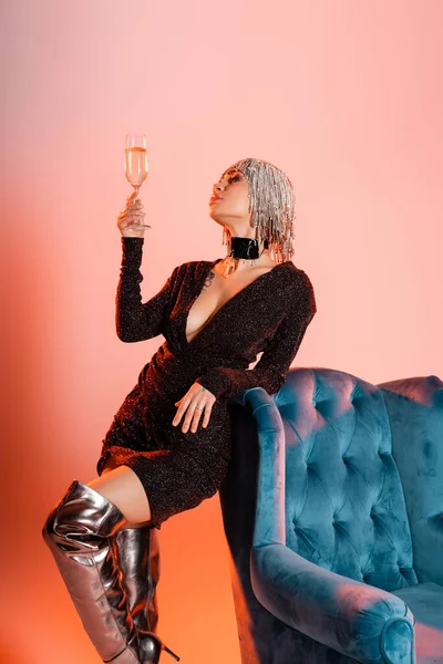 Mulher em peruca de prata e vestido lurex preto de pé com copo de champanhe perto de poltrona de veludo no fundo rosa e laranja — Fotografia de Stock