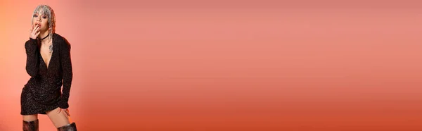 Femme passionnée en robe noire et coiffure métallique touchant les lèvres tout en se tenant debout sur fond rose corail, bannière — Photo de stock