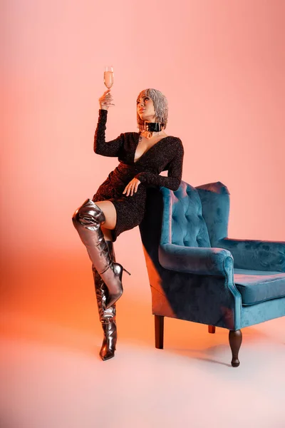 Pleine longueur de la femme en bottes longues et robe noire tenant champagne près du fauteuil sur fond rose et orange — Photo de stock