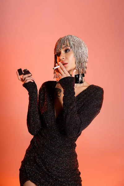 Verführerische Frau in metallischer Kopfbedeckung und schwarzem Lurexkleid, Zigarette rauchend auf korallenrosa Hintergrund — Stockfoto