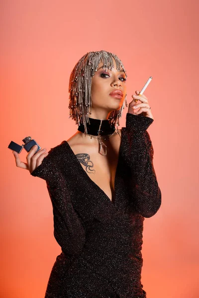 Femme à la mode en bijoux coiffure posant avec cigarette et briquet sur fond rose corail — Photo de stock