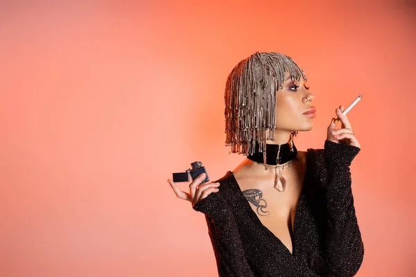 Tattooed woman in metallic wig and dress holding cigarette and looking away on pink and orange background — Stock Photo