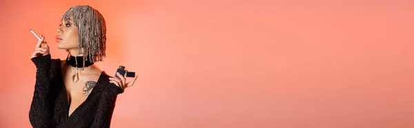 Femme tatouée sexy en robe noire et perruque argentée avec strass tenant cigarette et regardant loin sur fond rose corail, bannière — Photo de stock