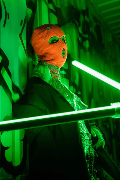 Sexy woman in orange balaclava and black blazer looking at camera near bright neon lamps and green wall with graffiti — Stock Photo