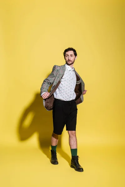 Longitud completa del modelo de moda en pantalones cortos tocando chaqueta sobre fondo amarillo con sombra - foto de stock