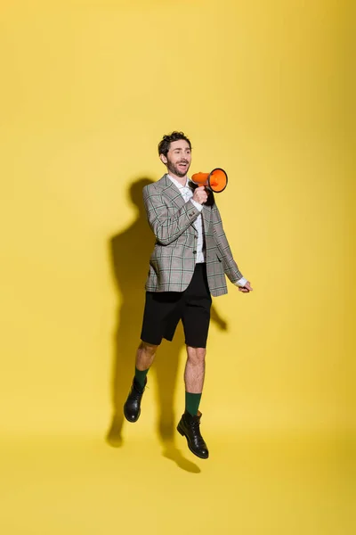 Homem alegre e elegante em shorts e jaqueta segurando alto-falante enquanto salta no fundo amarelo — Fotografia de Stock
